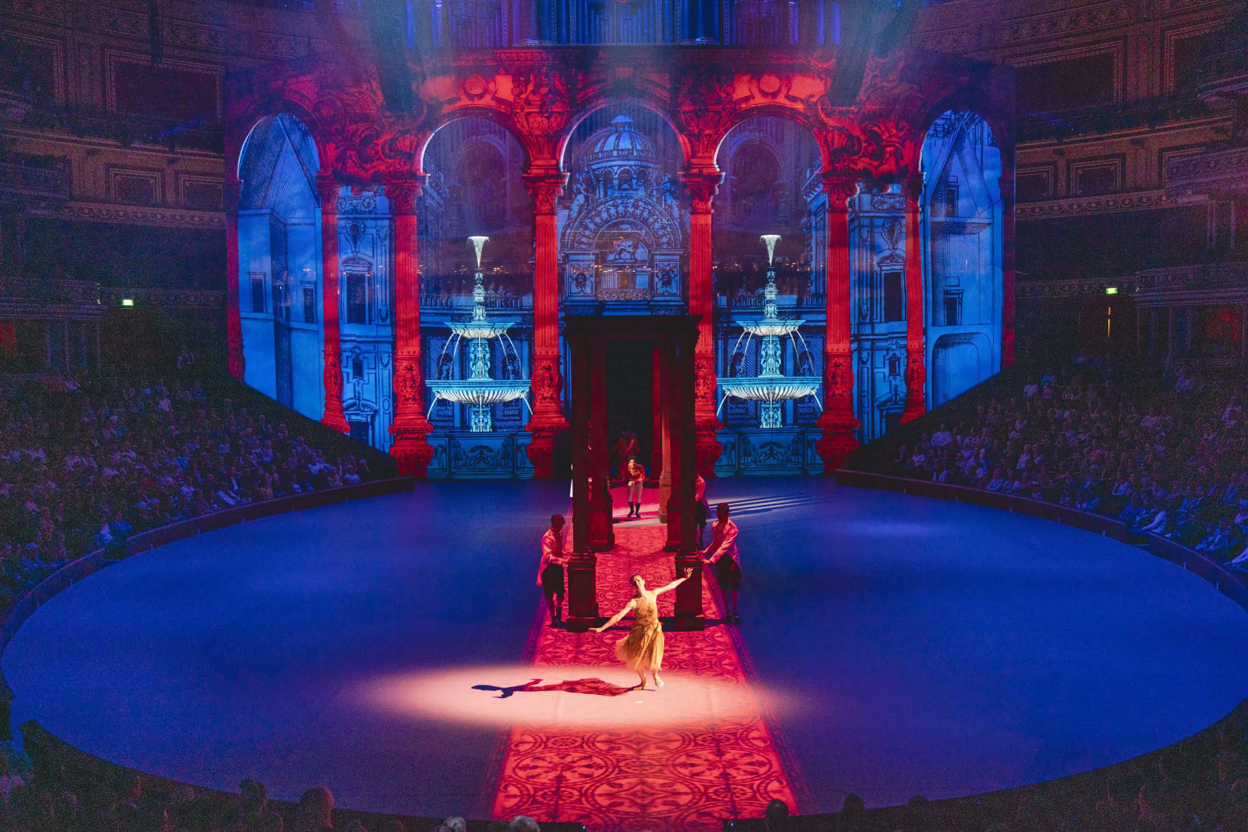 A scene from Christopher Wheeldon's ballet Cinderella in-the-round at the Royal Albert Hall, featuring immersive projection design. The stage is bathed in blue and red lighting, with digital projections of grand arches and fountains creating a striking, classical architectural backdrop. Dancers in period costumes perform under the atmospheric lighting, enhancing the theatrical experience.