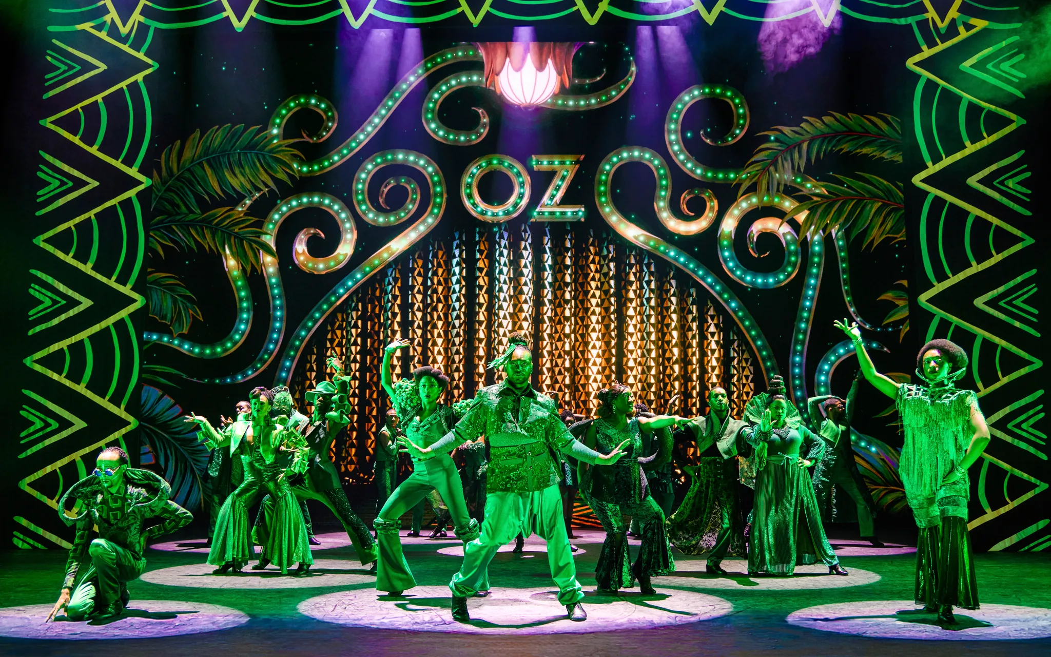A vibrant stage production of The Wiz, with performers in green costumes dancing under green lights. In the background, the word 'OZ' is illuminated in large, stylized green letters, creating a magical Emerald City atmosphere.
